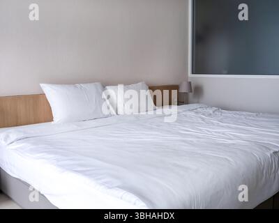 White clean bedroom in guest hotel room interior, white blanket with two pillows on simple wooden bed, near frosted glass partition and empty wall, to Stock Photo