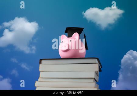 A pink piggy bank wearing a black graduation cap on top of a stack of books against a bright blue sky with clouds. Academic achievement Stock Photo