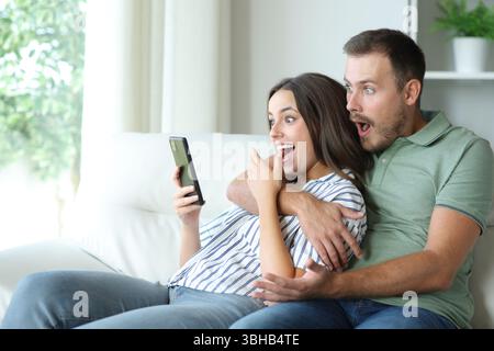 Surprised couple checking mobile phone content sitting on a couch at home Stock Photo
