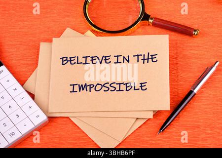 Motivational quote. BELIEVE IN THE IMPOSSIBLE text written on an envelope on a bright background, along with a magnifying glass, calculator, and pen. Stock Photo