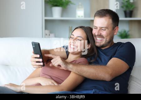 Happy couple using cellphone together sitting in a couch at home Stock Photo