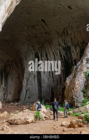 Group of hikers stands at massive cave entrance with towering vertical wall and rock layers. Stock Photo