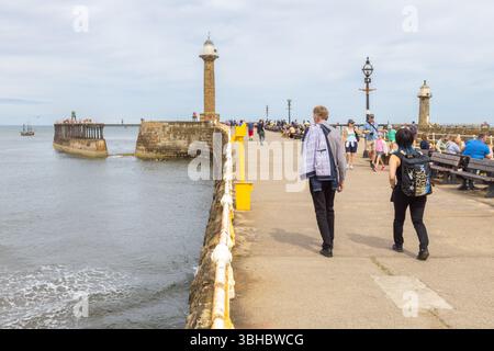 Tourists strolling along Whitby’s West Pier toward the lighthouses, with the HMS Endeavour replica sailing to the left against the sea and horizon. Stock Photo