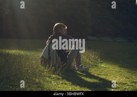 Man sitting on grass with backpack in a sunlit park Stock Photo