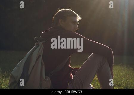 Young man relaxing on grass with backpack under warm sunlight Stock Photo