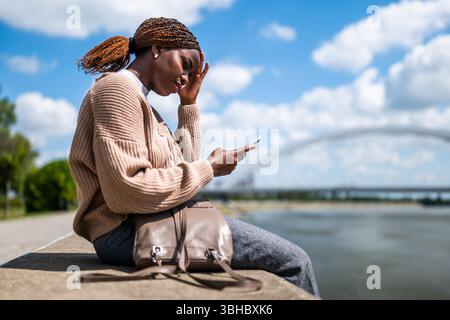 Young worried black woman is using smartphone. She is sitting by the river in the city. Stock Photo