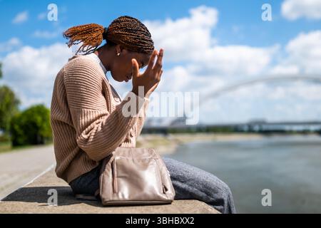 Portrait of displeased young black woman who is sitting by the river in the city. Stock Photo
