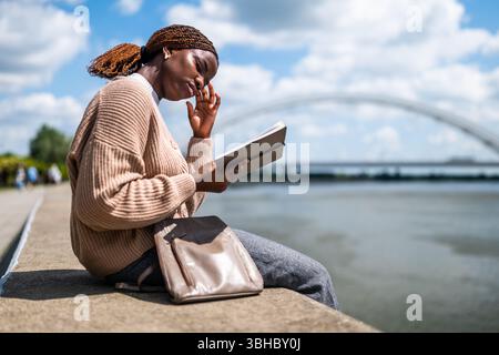 Young black woman is sitting by the river in the city. She is relaxing and reading book. Stock Photo