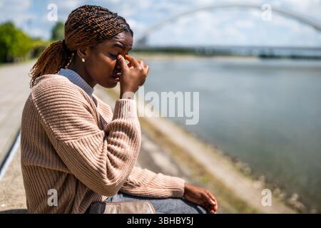 Young beautiful black woman is sitting by the river in the city. She is having headache. Stock Photo
