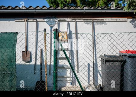 Garden tools and ladder hanging on white wall behind fence, concept of outdoor organization Stock Photo