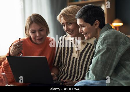 Three middle aged Caucasian women sitting closely together using laptop, smiling and interacting, demonstrating friendship and connection in casual indoor setting Stock Photo