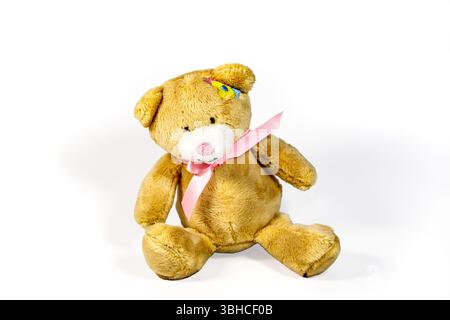 Cute brown teddy bear with a pink ribbon sitting on a white background, displaying a colorful patch on its head Stock Photo