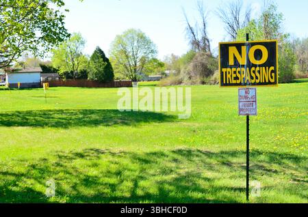 Series of No Trespassing signs across an open park with trees and houses in the background and a blue morning sky above, casting shadows. Stock Photo