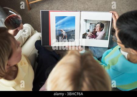 Three young adult Caucasian women sitting together viewing photo album, one holding glass of red wine, friends reminiscing and sharing memories while looking at printed photographs Stock Photo