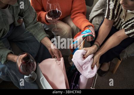 Three young adult women sitting together holding glasses of red wine and examining shopping bags and colorful scarves, hands and arms visible, showing friendship and leisure activity Stock Photo