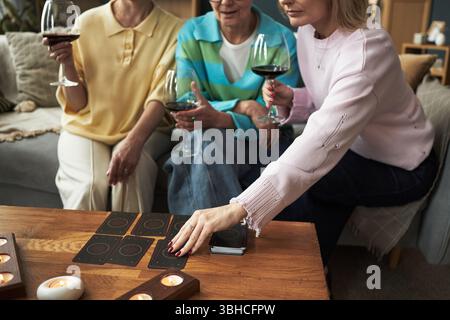 Three middle aged Caucasian women sitting together holding wine glasses, playing card game on wooden table, one woman reaching for card, candles burning in foreground Stock Photo