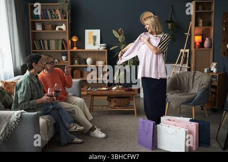 Three middle aged Caucasian women relaxing in living room, smiling and chatting while holding wine glasses, one woman standing and showing new clothing item from shopping bags Stock Photo