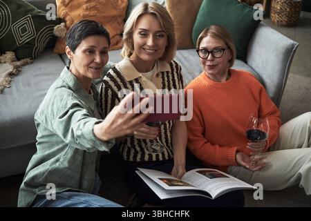Three middle aged Caucasian women sitting on floor taking selfie with smartphone, smiling and holding wine glasses, one woman holding open book Stock Photo