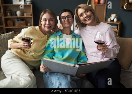 Three Caucasian women sitting closely on sofa, middle aged woman in glasses holding photo album, two young adult women smiling and holding wine glasses, enjoying time together Stock Photo