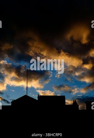 Silhouette of rooftops against a vibrant sunset sky. The sky is a mix of dark, shadowy areas and patches of bright orange and blue, creating a dramati Stock Photo