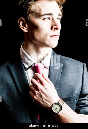 A young man in a suit adjusts his pink tie. He is wearing a watch with a unique face, and his expression is thoughtful. Stock Photo