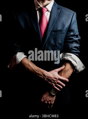 A man in a suit with a pink tie and his shirt sleeves rolled up. He is wearing a watch on his left wrist and is holding his right arm with his left ha Stock Photo