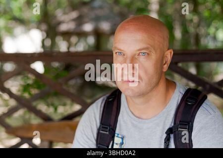 Man with a bald head and a backpack sits on a bench. He is wearing a gray shirt and a blue backpack Stock Photo