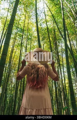 Back view of a woman with long wavy hair and a straw hat standing in a dense bamboo forest. She is wearing a light pink dress and looking up at the towering bamboo stalks surrounding her. The sunlight filters through the green canopy, creating a peaceful and immersive atmosphere. Captures the essence of nature exploration, mindful travel, and eco tourism in Asia.  Stock Photo