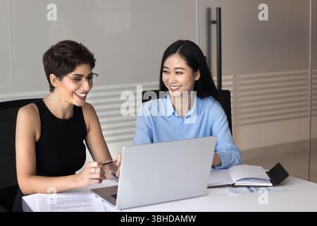 Two attractive multiethnic women colleagues sitting at desk with laptop Stock Photo