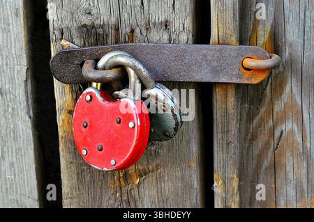 Close-up of an old red padlock on a weathered wooden door, secured with a rusty metal latch, symbolizing security and abandonment. Stock Photo