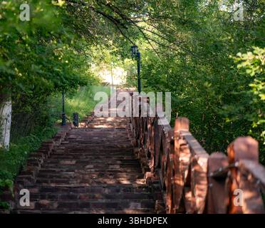 The photo shows a red brick staircase leading upward through a green forest. On the left are lampposts along the path, on the right — wooden railings. Stock Photo