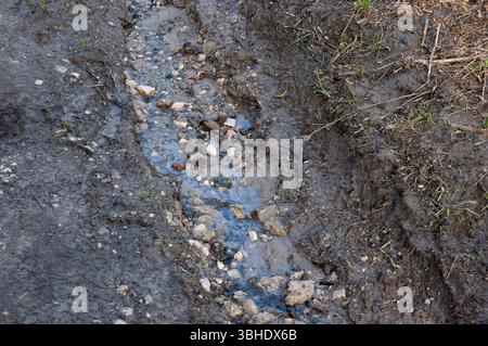 A muddy dirt road heavily eroded by a small stream. Water flows through a shallow channel filled with stones and broken bricks. Top-down view showing Stock Photo