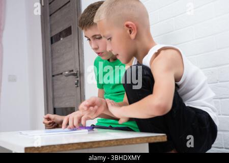 Two boys working together on a school project or game. One boy is crouching next to the table with the other who is sitting. They appear focused and a Stock Photo