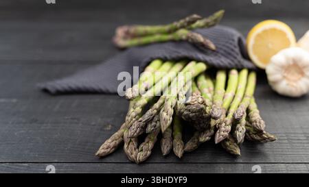 Close-up of green asparagus spears resting on dark wood with a deep blue kitchen towel, lemon and garlic in background Stock Photo