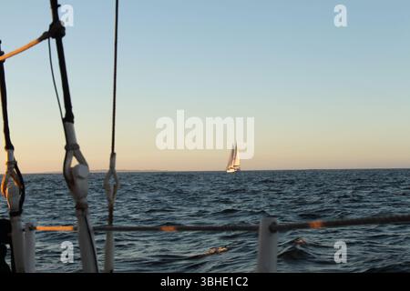 A lone sailboat glides across the horizon at sunset, seen from another vessel Stock Photo