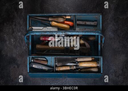 Fully opened vintage blue metal toolbox with various old tools in main and side compartments, on dark slate in studio setting Stock Photo