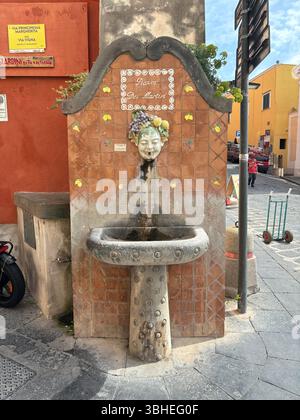 A rustic old stone water fountain with water flowing from a carved stone face’s mouth, blending natural charm and historic artistry outdoors. Stock Photo