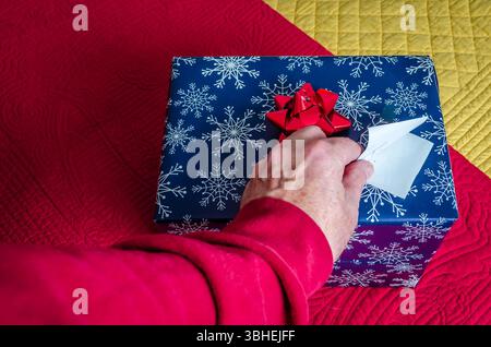 Top view of a man's hand opening the gift tag on a wrapped Christmas present sitting on red and yellow quilted bedspreads. Stock Photo