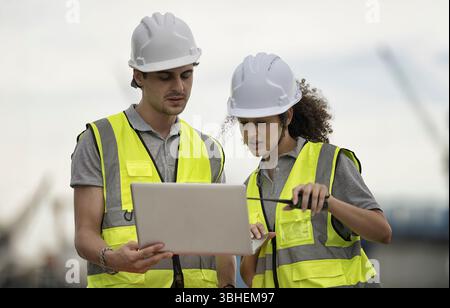 Two young workers in safety gear discuss information on a laptop at a construction site. They are focused, surrounded by industrial elements and equip Stock Photo
