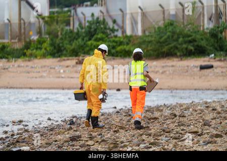 Two workers wearing protective gear walk along a rocky shoreline near an industrial area. They carry equipment and clipboards, focusing on an environm Stock Photo