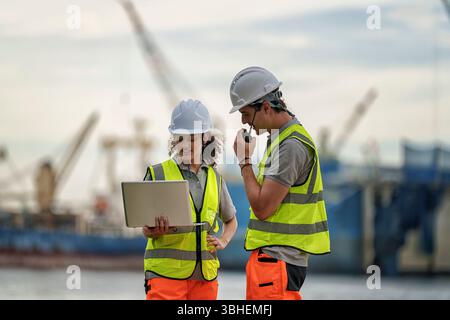 Two construction workers wearing safety gear are engaged in a discussion at a dock. They are looking at a laptop, planning or reviewing project detail Stock Photo
