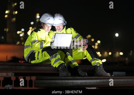 Two construction workers wearing high visibility gear are seated on a rail, focused on a laptop. They are collaborating on a project under bright indu Stock Photo