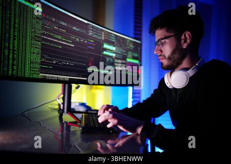 A young man sits at a sleek desk focused on coding, surrounded by glowing monitors displaying lines of code. The vibrant lighting enhances the tech at Stock Photo