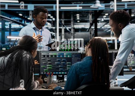 Engineers doing brainstorming, programming in server farm housing infrastructure storing datasets. Teamworking colleagues run code on PC to repair data center racks doing computational operations Stock Photo