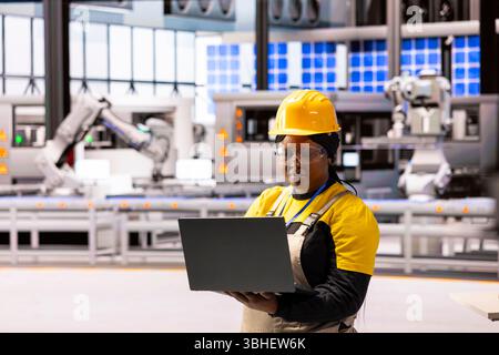 Female engineer in modern facility, overseeing operations and using laptop to ensure efficiency and safety. African american woman in factory, working on production of innovative devices. Stock Photo