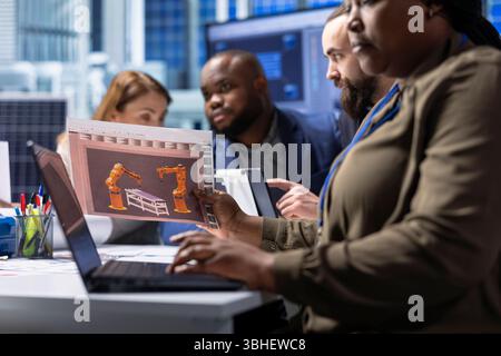Photovoltaics factory managers look over schematics, planning work on facility machines using laptop. Solar panels plant shareholders look over assembly lines blueprints, working on notebook Stock Photo