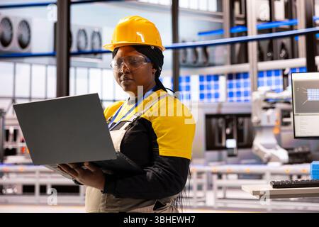 Black woman with industrial helmet using laptop to monitor and control equipment at sustainable energy production facility. African american engineer standing in factory and looking at device screen. Stock Photo