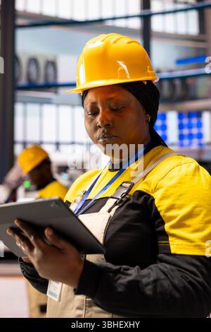Female engineer checking system performance on tablet in industrial manufacturing plant. Professional woman analyzes automation software data in smart factory, overseeing industry 4.0 production. Stock Photo
