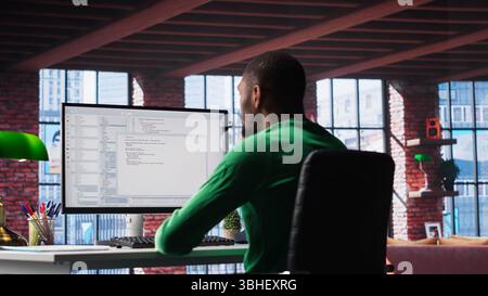Black IT developer at PC coding and creating an app software, implementing algorithms to fix errors. Engineer in his loft working with code language, optimizing app performance. Camera B. Stock Photo