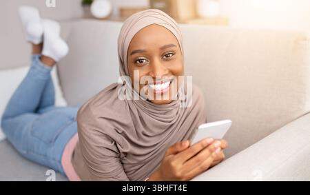 Online life. Smiling black woman in hijab lying on sofa with smartphone, chatting or browsing web at home Stock Photo